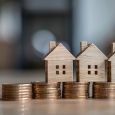 Three miniature wooden houses are positioned on stacks of gold coins, signifying the concept of homeownership and financial stability.