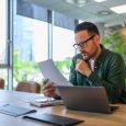 Focused young businessman in glasses reading document while working over computer at desk in office