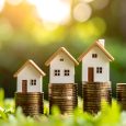 Miniature houses on increasing stacks of coins, set against a blurred green background