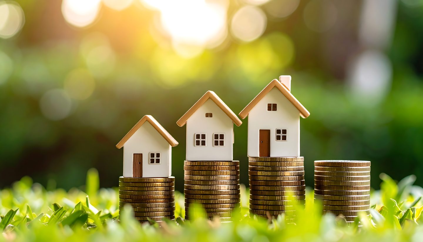 Miniature houses on increasing stacks of coins, set against a blurred green background