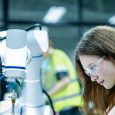 Two female robotics engineers in an R and D lab troubleshoot an artificial intelligence cobot. They are inspecting the robotic system hardware, sensors, and programming.