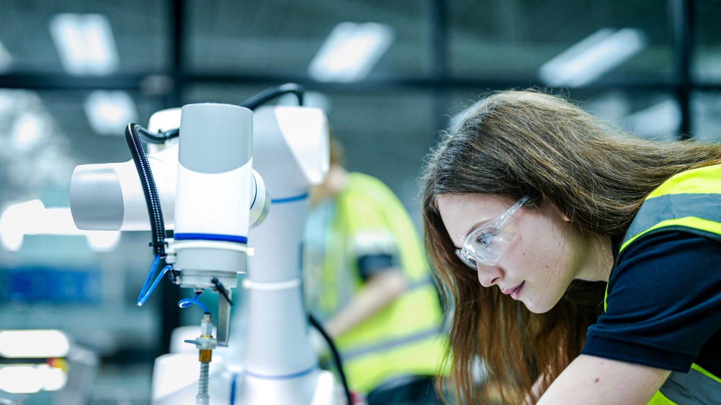 Two female robotics engineers in an R and D lab troubleshoot an artificial intelligence cobot. They are inspecting the robotic system hardware, sensors, and programming.